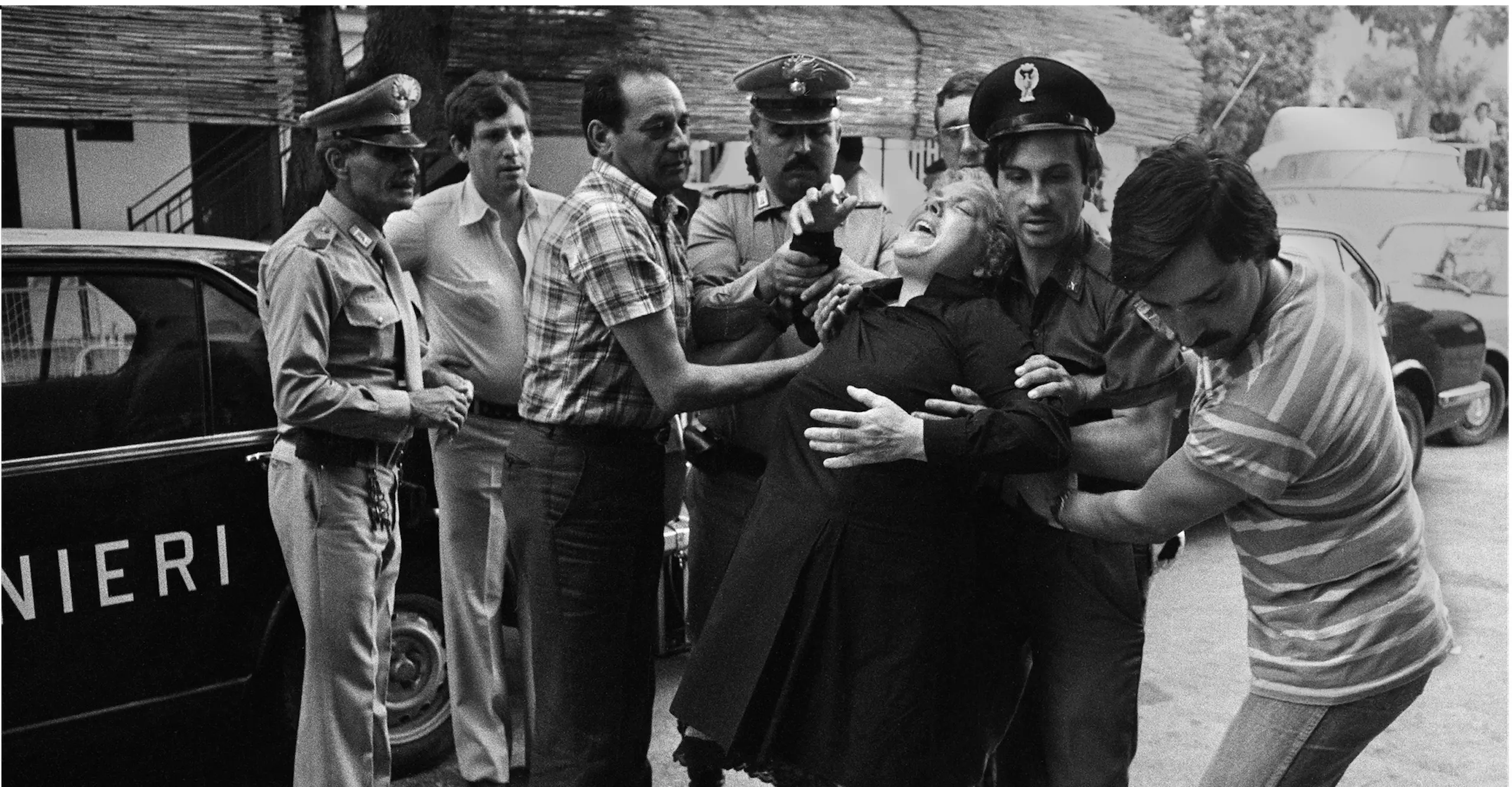 Black and white photograph of an inconsolable woman being held up by several police officers and a number of men. They try to calm her down.